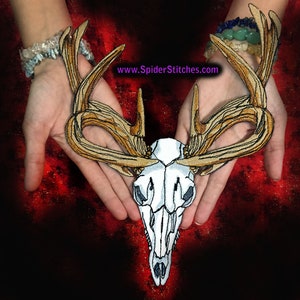 Könnte beinhalten: Ein weißer Hirschschädel mit großen, kunstvoll gestickten Geweih in Braun- und Goldtönen. Der Schädel wird in den Handflächen von zwei Händen gehalten, wobei der Text "www.SpiderStitches.com" oben im Bild sichtbar ist.