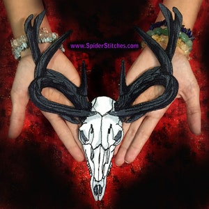 May include: A black and white embroidered deer skull patch with large antlers. The skull is white with black detailing, and the antlers are black. The patch is held in open hands against a red background. Text on the image reads: www.SpiderStitches.com.
