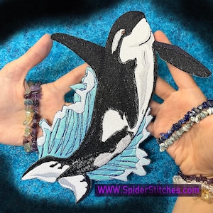 Può includere: Toppa ricamata in bianco e nero di un'orca che salta fuori dall'acqua. L'orca è circondata da acqua blu con schiuma bianca. La toppa ha il testo "WWW.SpiderStitches.com" in basso.