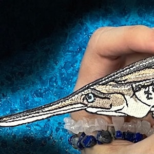 May include: A detailed embroidered patch of a longnose garfish, a type of fish with a long, thin snout and a streamlined body. The patch is beige and white with black details and is held in a hand.