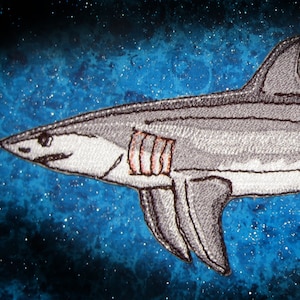 May include: A grey embroidered shark patch with a white belly and red gills. The shark is swimming to the right against a blue and white starry background.