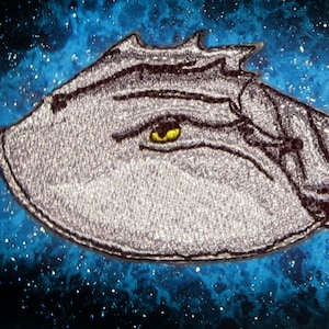 Może przedstawiać: Szara haftowana łatka w kształcie kraba podkowiastego z żółtym okiem. Łatka znajduje się na niebieskim tle z białymi gwiazdami.