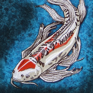 Könnte beinhalten: Ein weißer und roter Koi-Fisch mit schwarzen Markierungen ist auf einem blauen Hintergrund gestickt. Der Fisch hat einen langen, fließenden Schwanz und Flossen.