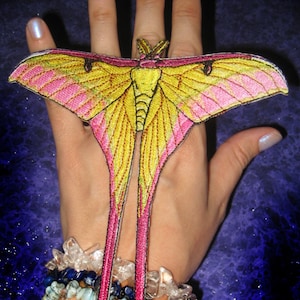 May include: A pink and yellow embroidered moth with long tails, attached to a person's wrist. The moth is a popular symbol of transformation and rebirth.