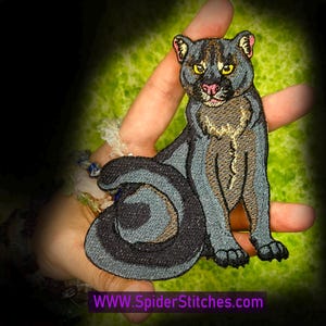 May include: An embroidered patch featuring a seated panther with dark grey and black fur, yellow eyes, and a pink nose. The design includes a long, curled tail and detailed stitching. The text WWW.SpiderStitches.com is printed at the bottom.