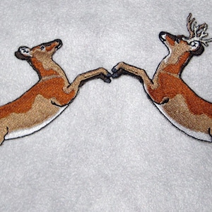 Puede incluir: Dos ciervos marrones bordados con detalles blancos saltan en direcciones opuestas. Los ciervos se miran con la cabeza echada hacia atrás.