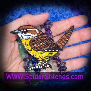 May include: Embroidered patch of a small bird with brown, yellow, and white details. The bird is perched on a strand of purple and green beads. The text "WWW.SpiderStitches.com" is displayed in a purple banner at the bottom of the image.