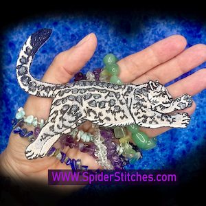May include: An embroidered patch featuring a stylized snow leopard in black and white, with a dark blue tail. The patch is held over a collection of colorful beads. The website address "WWW.SpiderStitches.com" is printed in purple.