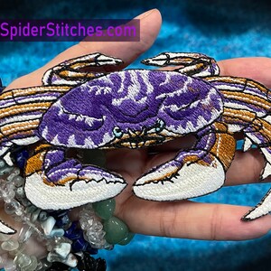 May include: Purple and gold embroidered crab patch with white details. The patch is held in a hand with a bracelet on the wrist.