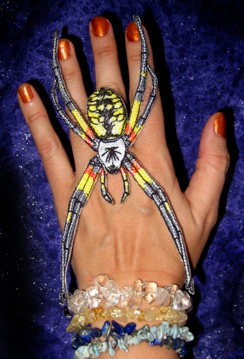 May include: A yellow and black spider with red markings is embroidered on a hand wearing a bracelet with multiple strands of clear, yellow, blue, and turquoise stones.