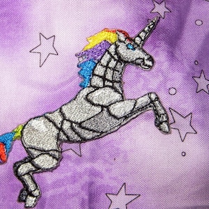 Può includere: Un unicorno argentato con criniera e coda arcobaleno è ricamato su uno sfondo di tessuto viola con stelle e una luna crescente.
