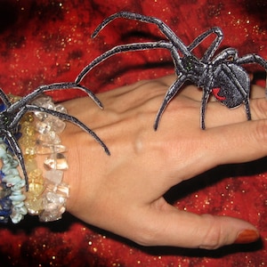 Puede incluir: Dos parches bordados de araña negra en la muñeca y la mano de una persona. Las arañas tienen marcas rojas en sus abdómenes.