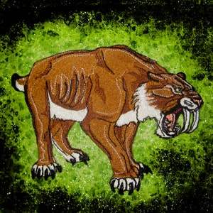 Può includere: Una toppa ricamata che raffigura una tigre dai denti a sciabola con grandi zanne, in marrone e bianco, su uno sfondo scintillante verde e nero.