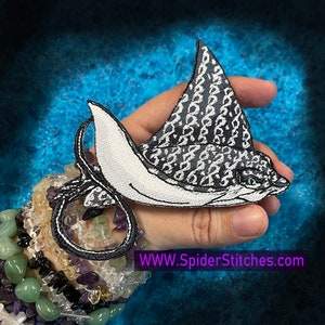 Peut inclure: Un patch brodé noir et blanc représentant une raie manta, avec un fond blanc et des taches noires. Le patch est tenu dans une main ornée d'une pile de bracelets. Le texte "WWW.SpiderStitches.com" est visible dans le coin inférieur droit de l'image.
