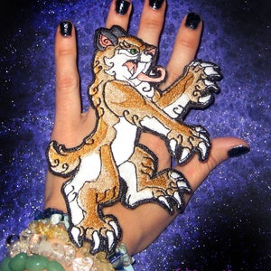 Może przedstawiać: Naszywka haftowana przedstawiająca ryczącego szablastozębnego kota z długim językiem, w kolorze brązowym i białym. Naszywka jest umieszczona na dłoni z czarnym lakierem do paznokci.