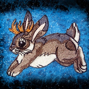 May include: A brown and gray embroidered rabbit with antlers, leaping against a blue starry background.