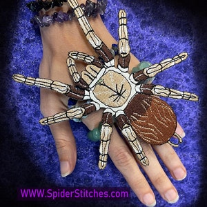 Puede incluir: Un parche de araña bordado en marrón y blanco con un patrón de rayas blancas y negras en el abdomen. El parche está en la mano de una persona, con un fondo azul.