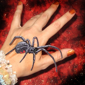 以下が含まれることがあります： 赤い砂時計のマークが付いた黒いクモのアップリケ。クモは長く細い脚と緑色の目をしています。背景はキラキラした濃い赤色です。