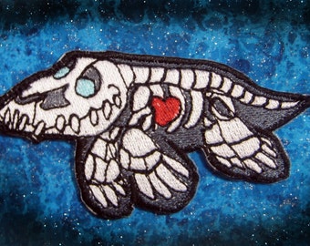 May include: Embroidered patch featuring a stylized skeleton of a creature with a red heart. The patch has a black border, white bones, and light blue eyes. The background is a blue and black galaxy design.