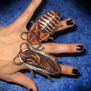 Puede incluir: Dos parches bordados para planchar de cucarachas marrones y blancas. Los parches tienen ojos rojos y están sobre un fondo azul.