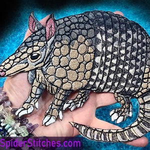 Puede incluir: Un parche bordado detallado de un armadillo, con una concha beige y marrón con acentos negros y blancos. El armadillo está representado en una pose de caminar con la cabeza girada hacia un lado. El parche mide aproximadamente 15 cm de largo y 10 cm de ancho.