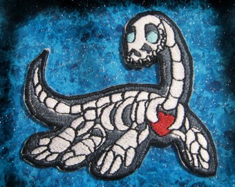 May include: Embroidered patch of a white skeleton sea serpent with a red heart on a blue sparkly background.