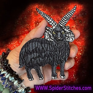 以下が含まれることがあります： 赤いキラキラした背景に、手の中に置かれた黒い刺繍の羊のワッペン。羊は大きく巻いた角を持ち、黄色い目をしています。羊の体は黒くふわふわしています。