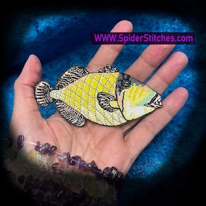 May include: Embroidered fish patch with yellow, black, and green details. The fish has a diamond pattern on its body and a detailed design. The patch is held in a hand. Text on the image reads: WWW.SpiderStitches.com.