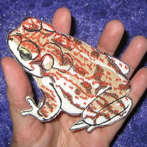 May include: A brown and white embroidered patch of a toad with red markings.
