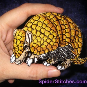 May include: A yellow and brown embroidered patch of an armadillo. The armadillo is curled up in a ball with its legs and tail visible. The patch is about 3 inches wide and 2 inches tall.
