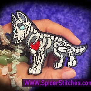 May include: Embroidered iron-on patch of a skeleton dog with a red heart in its chest. The dog is standing on all fours and has a white and gray color scheme. The patch is on a hand with a purple background.