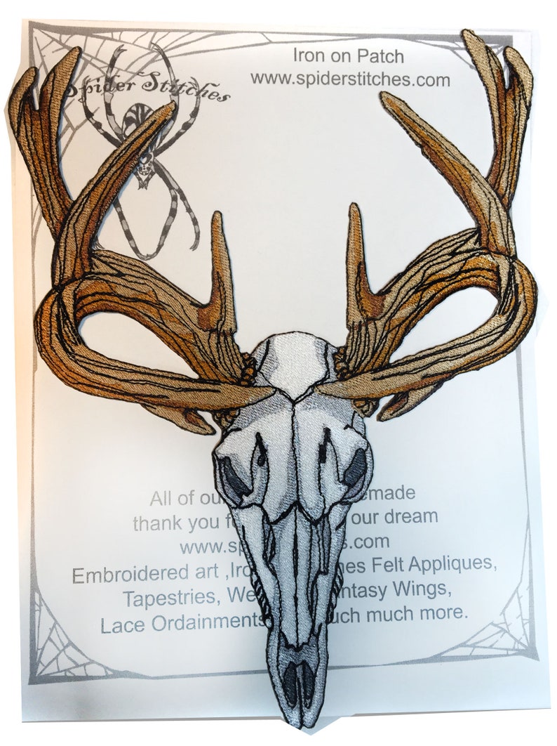 May include: Iron-on patch featuring a white embroidered deer skull with brown antlers. The patch is on a white background with text that reads "Iron on Patch" and "www.spiderstitches.com".