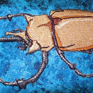 Könnte beinhalten: Ein gestickter Käfer mit großen Hörnern in Braun- und Schwarztönen. Der Käfer befindet sich auf einem blauen Hintergrund mit glitzernder Textur.