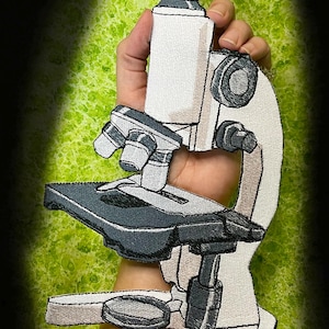 Puede incluir: Parche bordado de un microscopio en tonos blanco, gris y negro. El parche se sostiene sobre un fondo verde texturizado. La dirección del sitio web WWW.SpiderStitches.com es visible en la parte inferior.