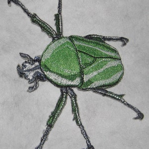 May include: Green and white embroidered beetle with six legs and a detailed design.