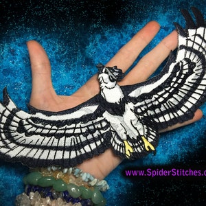 May include: A black and white embroidered patch of an osprey with its wings spread wide. The patch is on a hand against a blue background with white stars.
