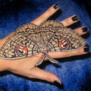 Könnte beinhalten: Ein brauner und schwarzer gestickter Falter mit roten Augen auf einer Hand mit schwarzem Nagellack.