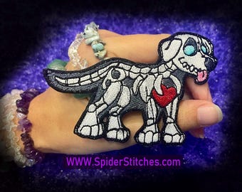 May include: A black and white embroidered patch of a skeleton dog with a red heart. The dog has a blue eye and a pink tongue. The patch is on a hand with a purple and white bracelet. The text "WWW.SpiderStitches.com" is at the bottom of the image.