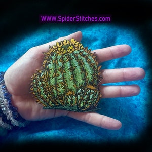 May include: A green and gold embroidered cactus patch with yellow flowers. The patch is on a hand, with the text "WWW.SpiderStitches.com" in the background.