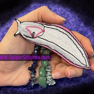 May include: White and pink embroidered iron-on patch of a sea slug. The patch is approximately 13 cm long and 5 cm wide. The patch is on a hand with a stack of bracelets. The text "WWW.SpiderStitches.com" is visible in the image.