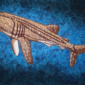 May include: A brown and white whale shark embroidered on a blue background. The shark is swimming to the right and has a long, slender body with a pointed snout. The background is a mottled blue with white flecks.