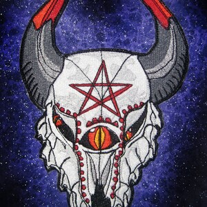 May include: Embroidered patch featuring a white skull with a red pentagram and a single red eye. The skull has horns and is set against a dark blue starry background. The text "spiderstitches.com" is visible at the top of the image.