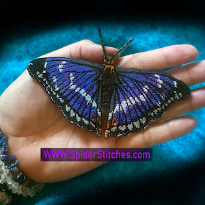 以下が含まれることがあります： 白のアクセントが付いた、紫と黒の刺繍された蝶のワッペン。ワッペンは、青と白のビーズのブレスレットをした手に置かれています。