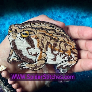 Op de afbeelding: Gestikt patch van een bruine en witte pad met gele ogen. De pad zit op een hand met een armband om de pols. De tekst "WWW.SpiderStitches.com" is zichtbaar onder de pad.