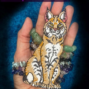 May include: Embroidered patch featuring a detailed bobcat design with tan, black, and white fur, and pink ear accents. The bobcat is in a seated position. The website address WWW.SpiderStitches.com is visible at the top.