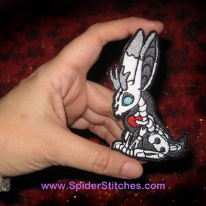 May include: A black and white embroidered patch of a skeletal bunny holding a red heart. The bunny has large, white ears and blue eyes. The patch is about 3 inches tall.