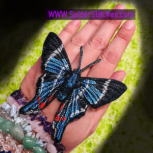 May include: A blue and black butterfly embroidered patch with white accents. The butterfly has red accents on the wings. The patch is on a hand.