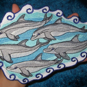 May include: Embroidered patch featuring five grey dolphins swimming in a blue and white wave pattern. The dolphins are stylised and have a whimsical look.