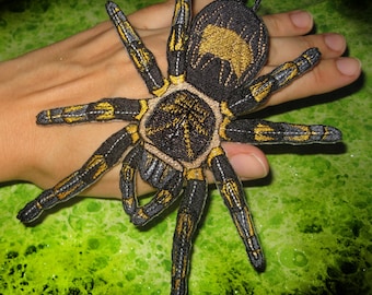 May include: Embroidered spider patch featuring a black body with gold and gray accents. The spider has eight legs and a detailed design. The patch is likely for clothing or accessories.