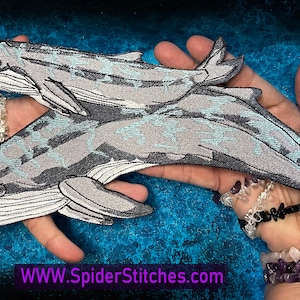 Può includere: Due toppe di balena ricamate, una più grande dell'altra, sono tenute nelle mani di una persona. Le balene sono grigie con un motivo blu chiaro e presentano punti di cucito dettagliati. Le toppe sono in vendita sul sito web www.spiderstitches.com.